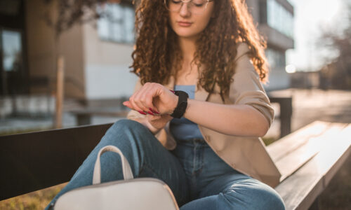 young woman with glasses looking at her smartwatch on a public outdoor bench