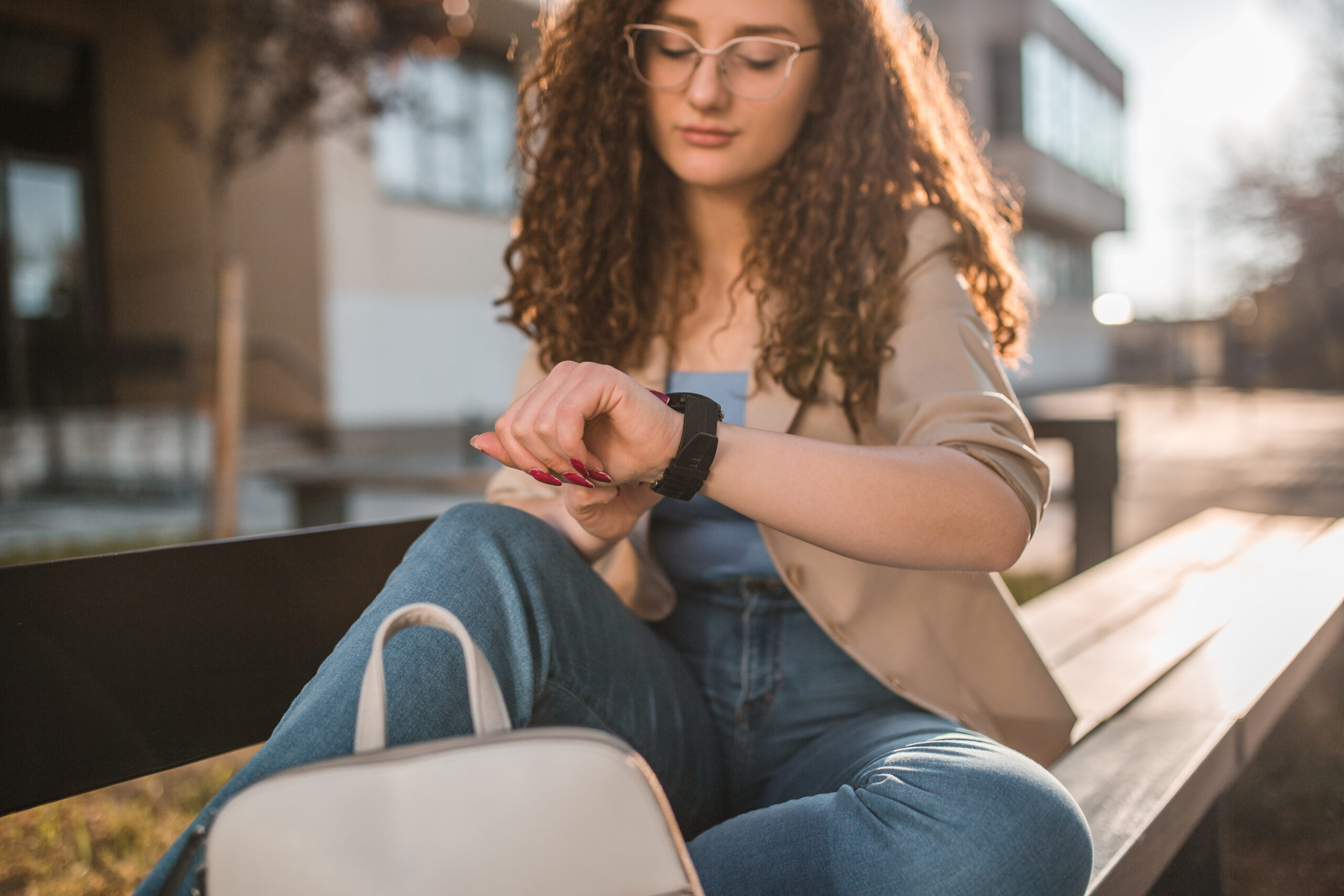 young woman with glasses looking at her smartwatch on a public outdoor bench