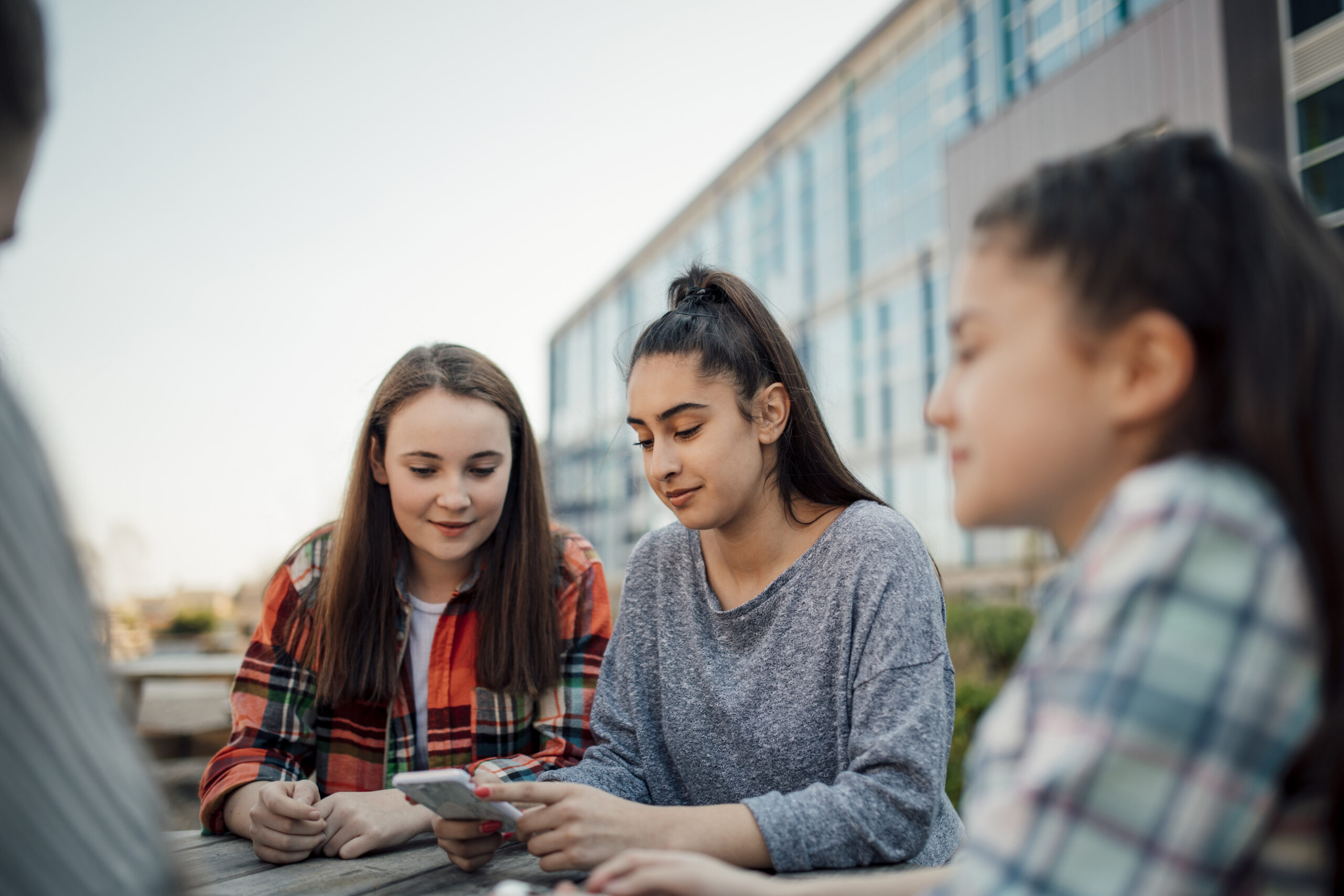 A group of schoolgirls sit talking at a table outside of school looking at their smartphone