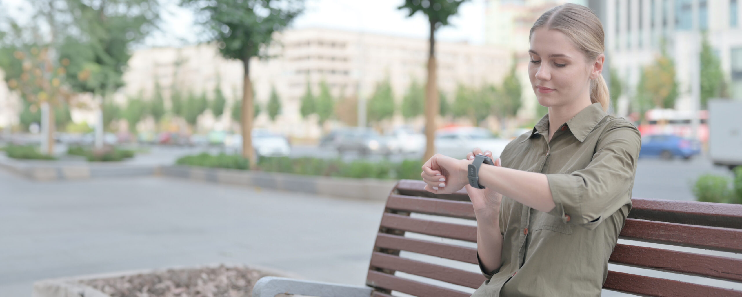 young woman with blonde hair sat on an outdoor bench looking at her smartphone watch